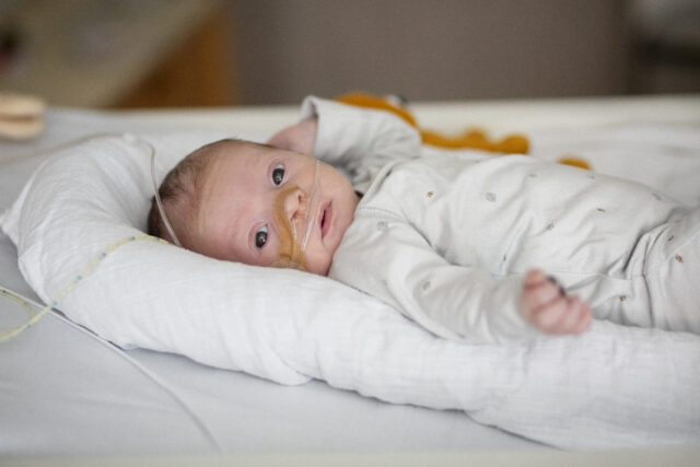 Pasgeboren baby ligt op een ziekenhuisbed met een zuurstofslangetje in de neus, rustig kijkend in de camera. De baby draagt een lichtgrijs rompertje en ligt op een zacht kussen.